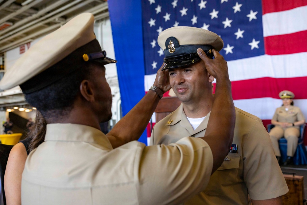 A U.S. Navy promotion or advancement ceremony is taking place, with one sailor placing a chief petty officer cover (hat) on another sailor's head. The ceremony appears to be held aboard a naval vessel or facility, with a large American flag displayed prominently in the background. This is consistent with a traditional Navy Chief Petty Officer pinning/advancement ceremony.