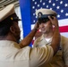 A U.S. Navy promotion or advancement ceremony is taking place, with one sailor placing a chief petty officer cover (hat) on another sailor's head. The ceremony appears to be held aboard a naval vessel or facility, with a large American flag displayed prominently in the background. This is consistent with a traditional Navy Chief Petty Officer pinning/advancement ceremony.