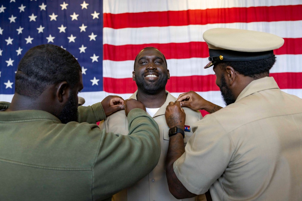A military promotion ceremony is taking place in front of an American flag backdrop. Two service members are pinning new rank insignia onto the collar of a smiling central figure, who appears to be receiving a promotion. The joyful expression of the central individual suggests this is a significant and celebratory milestone.