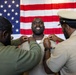 A military promotion ceremony is taking place in front of an American flag backdrop. Two service members are pinning new rank insignia onto the collar of a smiling central figure, who appears to be receiving a promotion. The joyful expression of the central individual suggests this is a significant and celebratory milestone.