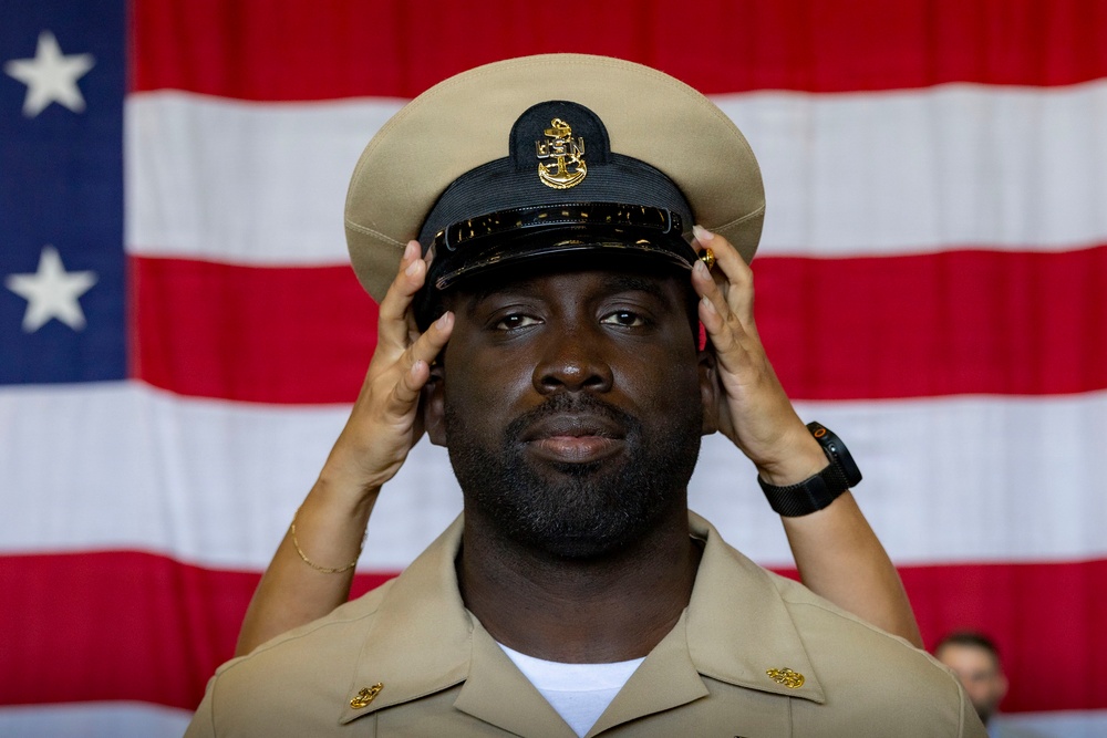 A U.S. Navy sailor in khaki service uniform is having a chief petty officer combination cover (cap) placed on his head, likely during a chief petty officer pinning or advancement ceremony. An American flag is prominently displayed in the background. The moment appears to be a formal military milestone, with another person's hands visible placing the cover on his head.