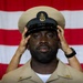 A U.S. Navy sailor in khaki service uniform is having a chief petty officer combination cover (cap) placed on his head, likely during a chief petty officer pinning or advancement ceremony. An American flag is prominently displayed in the background. The moment appears to be a formal military milestone, with another person's hands visible placing the cover on his head.