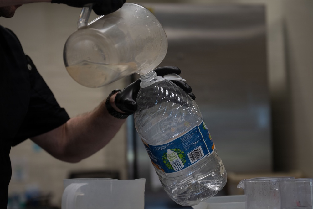 A person wearing black gloves and a black shirt is pouring liquid from a clear plastic measuring pitcher into a large plastic water jug. The large bottle has a blue label with recycling-themed graphics and a barcode visible. The setting appears to be a commercial or institutional kitchen environment.