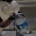 A person wearing black gloves and a black shirt is pouring liquid from a clear plastic measuring pitcher into a large plastic water jug. The large bottle has a blue label with recycling-themed graphics and a barcode visible. The setting appears to be a commercial or institutional kitchen environment.