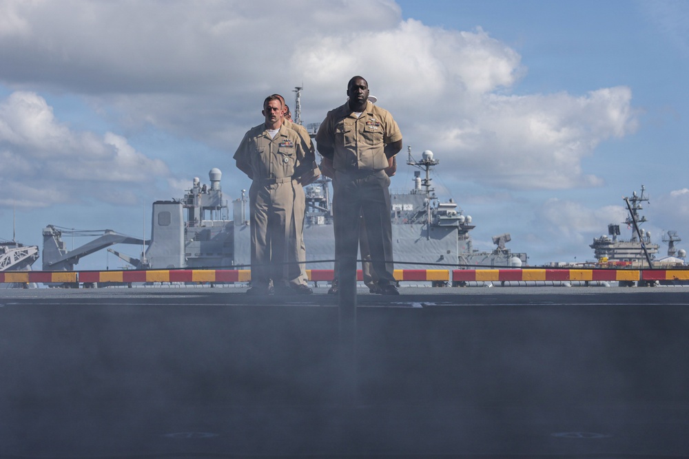 Two U.S. Navy personnel in tan/khaki service uniforms stand on what appears to be the flight deck or upper deck of a naval vessel. Behind them, at least two additional warships are visible in the background, suggesting a naval formation or port setting. The low camera angle creates a dramatic perspective with the figures silhouetted against a partly cloudy sky.