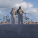 Two U.S. Navy personnel in tan/khaki service uniforms stand on what appears to be the flight deck or upper deck of a naval vessel. Behind them, at least two additional warships are visible in the background, suggesting a naval formation or port setting. The low camera angle creates a dramatic perspective with the figures silhouetted against a partly cloudy sky.