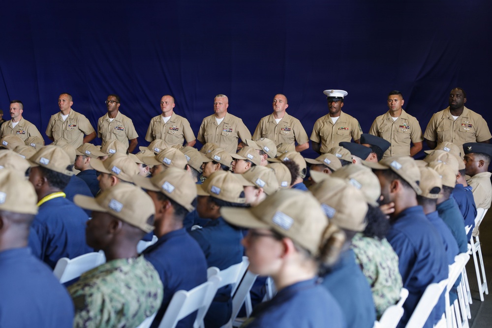 A formal military ceremony or gathering featuring uniformed U.S. Navy personnel standing in a row at the back, with recruits or junior personnel seated in rows wearing navy blue shirts and tan campaign-style hats. The backdrop is a large blue drape or curtain, suggesting an indoor or covered venue. One individual in the standing row wears a white cover (cap), consistent with U.S. Marine Corps dress uniform conventions.