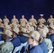 A formal military ceremony or gathering featuring uniformed U.S. Navy personnel standing in a row at the back, with recruits or junior personnel seated in rows wearing navy blue shirts and tan campaign-style hats. The backdrop is a large blue drape or curtain, suggesting an indoor or covered venue. One individual in the standing row wears a white cover (cap), consistent with U.S. Marine Corps dress uniform conventions.
