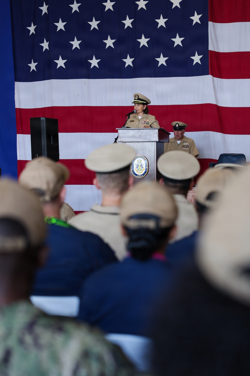 A U.S. Navy officer in service khaki uniform is speaking at a podium during what appears to be a formal military ceremony. A large American flag serves as the backdrop, and an audience of uniformed Navy personnel is seated facing the speaker. A second uniformed officer is visible seated behind and to the right of the podium.