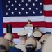 A U.S. Navy officer in service khaki uniform is speaking at a podium during what appears to be a formal military ceremony. A large American flag serves as the backdrop, and an audience of uniformed Navy personnel is seated facing the speaker. A second uniformed officer is visible seated behind and to the right of the podium.