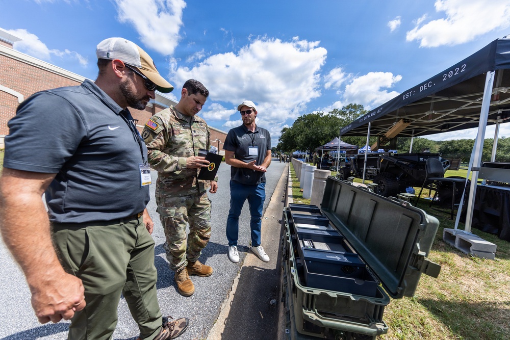 Three individuals — one in military camouflage uniform and two in civilian attire — are gathered outdoors examining a small device or item at what appears to be a military technology or equipment exposition. Open hard-shell tactical cases are visible in the foreground, and vendor tents with equipment displays line the background. The event appears to be a defense or military technology demonstration event, with a tent canopy marked 'EST: DEC. 2022' visible on the right.