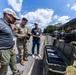 Three individuals — one in military camouflage uniform and two in civilian attire — are gathered outdoors examining a small device or item at what appears to be a military technology or equipment exposition. Open hard-shell tactical cases are visible in the foreground, and vendor tents with equipment displays line the background. The event appears to be a defense or military technology demonstration event, with a tent canopy marked 'EST: DEC. 2022' visible on the right.