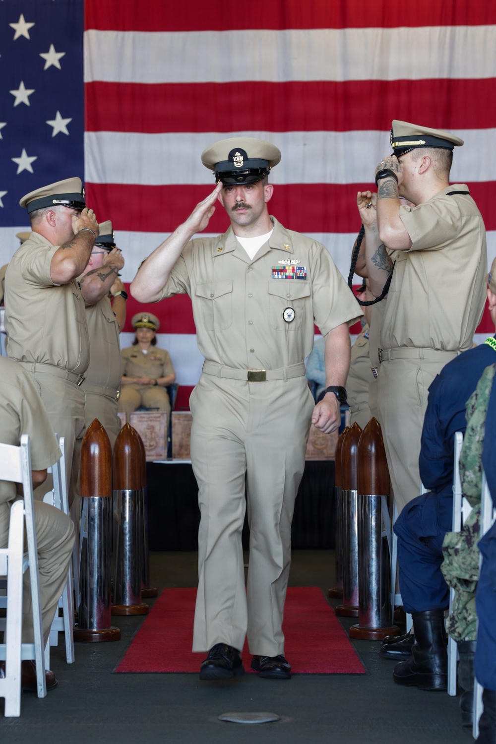 A U.S. Navy chief petty officer in khaki service uniform walks down a red carpet aisle while rendering a salute during a formal military ceremony. Flanking personnel on both sides also salute, and a large American flag serves as the backdrop. The setting includes decorative wooden and chrome bollards lining the aisle, suggesting a chief petty officer initiation or promotion ceremony.