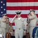 A U.S. Navy chief petty officer in khaki service uniform walks down a red carpet aisle while rendering a salute during a formal military ceremony. Flanking personnel on both sides also salute, and a large American flag serves as the backdrop. The setting includes decorative wooden and chrome bollards lining the aisle, suggesting a chief petty officer initiation or promotion ceremony.