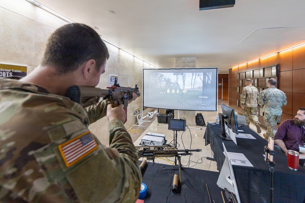 A U.S. Army soldier in MultiCam camouflage uniform is using a simulated weapons training system, aiming a rifle-shaped simulator at a projected screen displaying a virtual shooting range. The setup appears to be a demonstration or exhibition of a 'Virtual Trainer COTS' (Commercial Off-The-Shelf) marksmanship simulation system. Several other military personnel and a civilian operator are present in what appears to be an indoor facility or conference space.