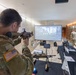 A U.S. Army soldier in MultiCam camouflage uniform is using a simulated weapons training system, aiming a rifle-shaped simulator at a projected screen displaying a virtual shooting range. The setup appears to be a demonstration or exhibition of a 'Virtual Trainer COTS' (Commercial Off-The-Shelf) marksmanship simulation system. Several other military personnel and a civilian operator are present in what appears to be an indoor facility or conference space.
