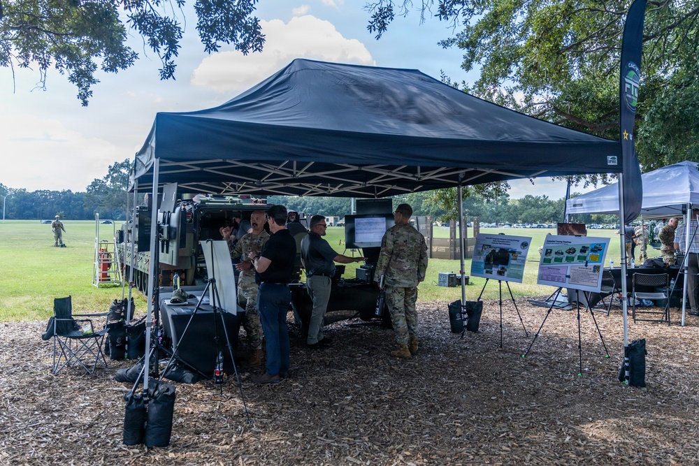 An outdoor military technology demonstration or expo event taking place on a grassy field under large canopy tents. Military personnel in camouflage uniforms and civilians are gathered around a military vehicle with mounted equipment and display screens. Informational poster boards are visible to the right side of the setup.