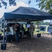 An outdoor military technology demonstration or expo event taking place on a grassy field under large canopy tents. Military personnel in camouflage uniforms and civilians are gathered around a military vehicle with mounted equipment and display screens. Informational poster boards are visible to the right side of the setup.
