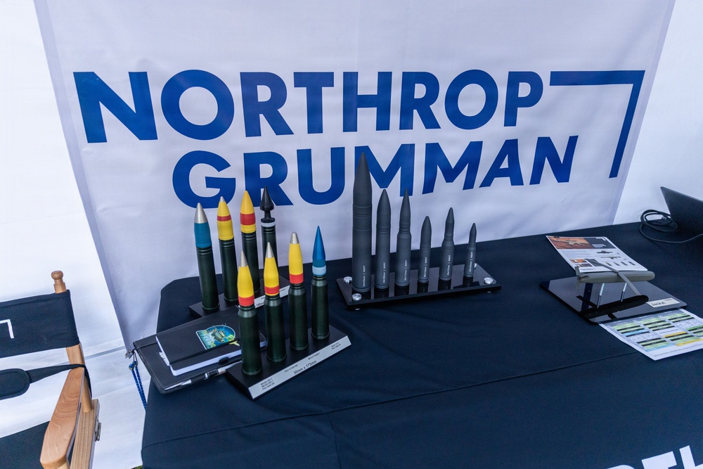 A Northrop Grumman trade show or exhibition booth displaying scale models or inert demonstration rounds of various munitions and projectiles on a navy blue draped table. The backdrop features the company's prominent blue-and-white logo banner. The display appears to be a professional defense industry showcase of ammunition and ordnance product lines.