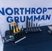 A Northrop Grumman trade show or exhibition booth displaying scale models or inert demonstration rounds of various munitions and projectiles on a navy blue draped table. The backdrop features the company's prominent blue-and-white logo banner. The display appears to be a professional defense industry showcase of ammunition and ordnance product lines.