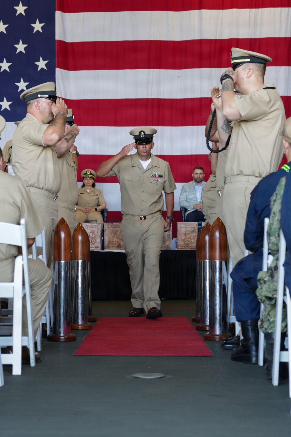A U.S. Navy military ceremony is taking place, likely a Chief Petty Officer pinning or advancement ceremony. A sailor in khaki service uniform walks down a red carpet aisle flanked by large decorative shell casings while fellow sailors render salutes on both sides. A large American flag serves as the backdrop, and seated observers including uniformed personnel and a civilian are visible in the background.