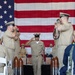 A U.S. Navy military ceremony is taking place, likely a Chief Petty Officer pinning or advancement ceremony. A sailor in khaki service uniform walks down a red carpet aisle flanked by large decorative shell casings while fellow sailors render salutes on both sides. A large American flag serves as the backdrop, and seated observers including uniformed personnel and a civilian are visible in the background.