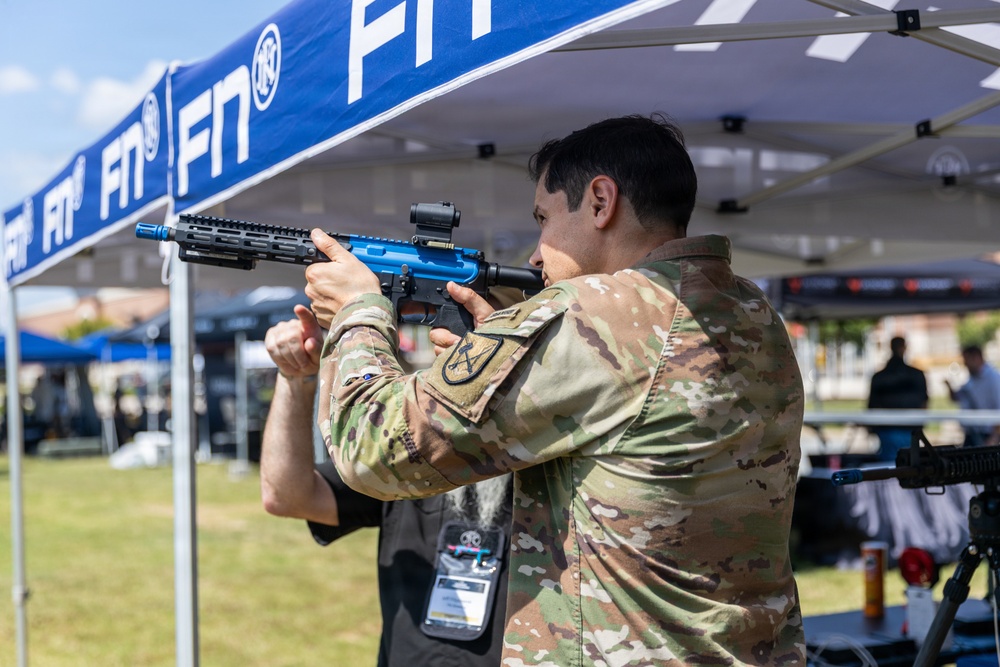 A person in military camouflage (OCP/MultiCam) uniform is aiming a blue-finished AR-style pistol/short-barreled rifle at an outdoor FN (Fabrique Nationale) branded exhibition or trade show event. The firearm appears to be a demonstration or display model, indicated by its distinctive blue anodized finish. The setting suggests a military or defense industry expo with vendor tents and other attendees visible in the background.
