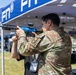 A person in military camouflage (OCP/MultiCam) uniform is aiming a blue-finished AR-style pistol/short-barreled rifle at an outdoor FN (Fabrique Nationale) branded exhibition or trade show event. The firearm appears to be a demonstration or display model, indicated by its distinctive blue anodized finish. The setting suggests a military or defense industry expo with vendor tents and other attendees visible in the background.