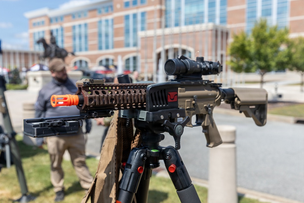 A rifle-style firearm mounted on a bipod/tripod is displayed in the foreground in sharp focus, with an orange muzzle device, optic scope, and attached accessory module visible. The setting appears to be an outdoor public area near a large brick building, suggesting a demonstration, exhibition, or display event. A blurred individual stands in the background near what appears to be an equestrian statue.