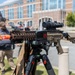 A rifle-style firearm mounted on a bipod/tripod is displayed in the foreground in sharp focus, with an orange muzzle device, optic scope, and attached accessory module visible. The setting appears to be an outdoor public area near a large brick building, suggesting a demonstration, exhibition, or display event. A blurred individual stands in the background near what appears to be an equestrian statue.