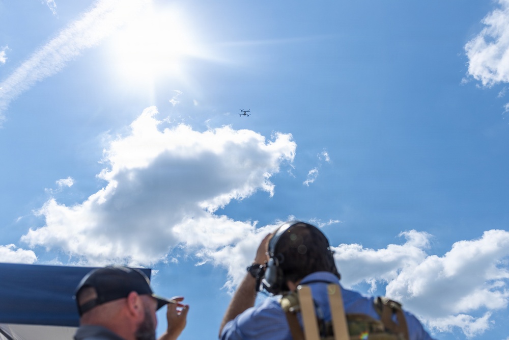 Two individuals are looking upward at a small drone or quadcopter visible in the sky. One person is wearing a baseball cap and appears to be speaking or gesturing, while the other is wearing headphones and a tactical vest and appears to be photographing or observing the aircraft. The scene suggests a drone operation or monitoring activity conducted outdoors under a bright, partly cloudy sky.