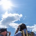 Two individuals are looking upward at a small drone or quadcopter visible in the sky. One person is wearing a baseball cap and appears to be speaking or gesturing, while the other is wearing headphones and a tactical vest and appears to be photographing or observing the aircraft. The scene suggests a drone operation or monitoring activity conducted outdoors under a bright, partly cloudy sky.
