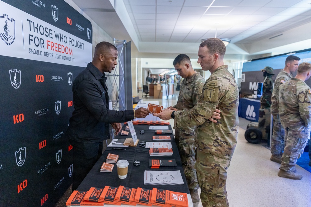 A civilian representative from 'Kingdom Operatives' (KO) is staffing an informational booth at what appears to be a military expo or resource fair, handing materials to uniformed soldiers in OCP (Operational Camouflage Pattern) uniforms. The booth displays the slogan 'Those Who Fought for Freedom Should Never Live Without It' and features orange-covered books titled 'Weapons of Mass Deception' on the table. The setting appears to be inside a military installation building with multiple vendor booths visible in the background.
