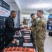 A civilian representative from 'Kingdom Operatives' (KO) is staffing an informational booth at what appears to be a military expo or resource fair, handing materials to uniformed soldiers in OCP (Operational Camouflage Pattern) uniforms. The booth displays the slogan 'Those Who Fought for Freedom Should Never Live Without It' and features orange-covered books titled 'Weapons of Mass Deception' on the table. The setting appears to be inside a military installation building with multiple vendor booths visible in the background.
