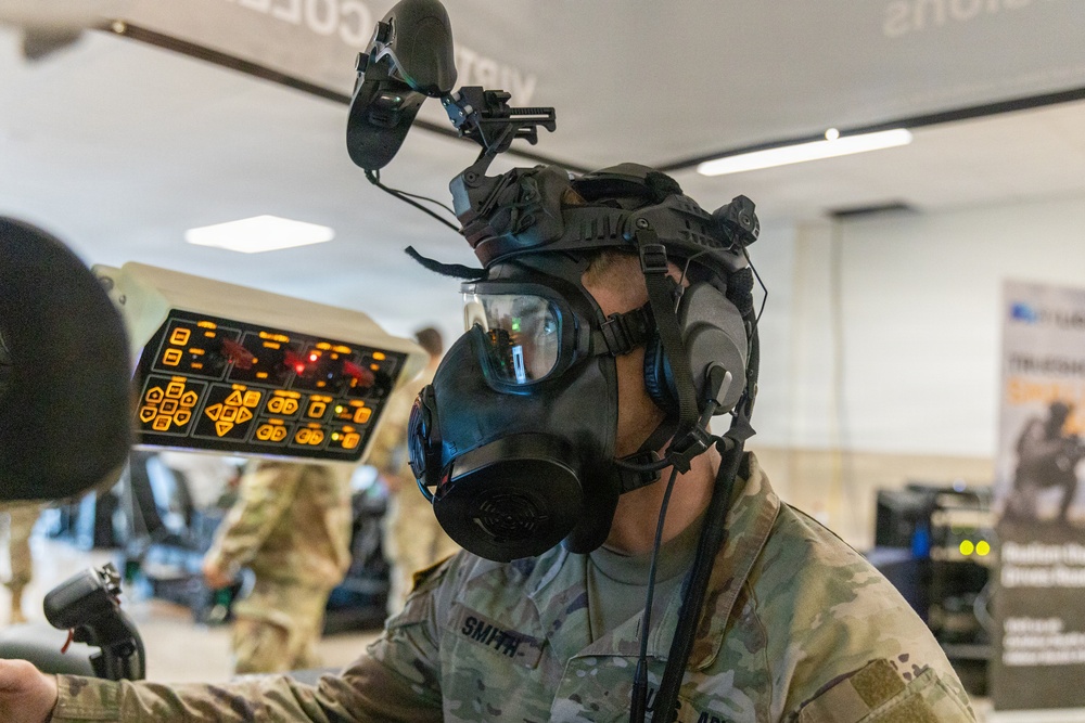 A military personnel member identified by the name tape 'SMITH' is wearing a full gas mask integrated with advanced tactical head-mounted equipment, including what appears to be night vision or sensor mounts. The setting appears to be an indoor military technology exhibition or demonstration event, with other uniformed personnel and display equipment visible in the background. The foreground shows a control panel with illuminated orange buttons and symbols.