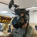 A military personnel member identified by the name tape 'SMITH' is wearing a full gas mask integrated with advanced tactical head-mounted equipment, including what appears to be night vision or sensor mounts. The setting appears to be an indoor military technology exhibition or demonstration event, with other uniformed personnel and display equipment visible in the background. The foreground shows a control panel with illuminated orange buttons and symbols.
