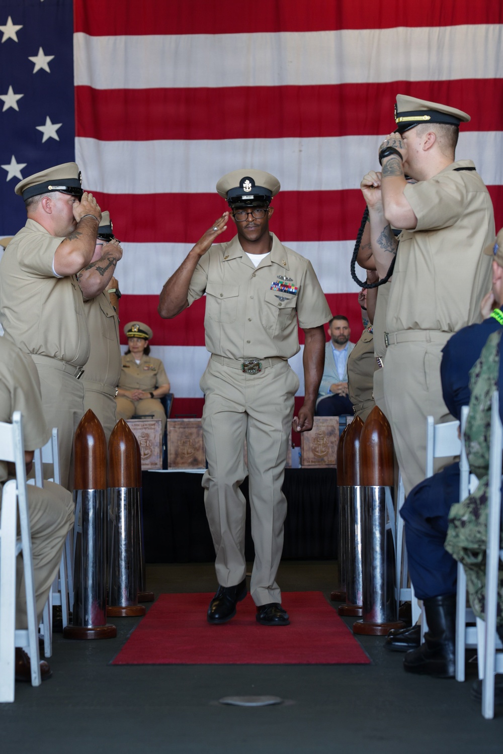 A U.S. Navy Chief Petty Officer walks down a red carpet aisle during what appears to be a Chief Petty Officer pinning or retirement ceremony. He is being saluted by fellow sailors on both sides as he passes between decorative shell casings lining the aisle. A large American flag serves as the backdrop for the formal military ceremony.