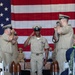 A U.S. Navy Chief Petty Officer walks down a red carpet aisle during what appears to be a Chief Petty Officer pinning or retirement ceremony. He is being saluted by fellow sailors on both sides as he passes between decorative shell casings lining the aisle. A large American flag serves as the backdrop for the formal military ceremony.