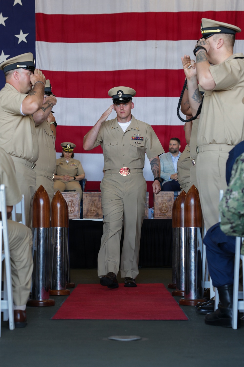 A U.S. Navy Chief Petty Officer induction ceremony is taking place, with a newly pinned Chief walking down a red carpet aisle while flanked by senior enlisted personnel rendering salutes. The ceremony is held indoors in front of a large American flag backdrop, with decorative wooden bullet/shell casings lining the aisle. The central figure is saluting while walking through a traditional 'gauntlet' formation.