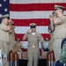 A U.S. Navy Chief Petty Officer induction ceremony is taking place, with a newly pinned Chief walking down a red carpet aisle while flanked by senior enlisted personnel rendering salutes. The ceremony is held indoors in front of a large American flag backdrop, with decorative wooden bullet/shell casings lining the aisle. The central figure is saluting while walking through a traditional 'gauntlet' formation.