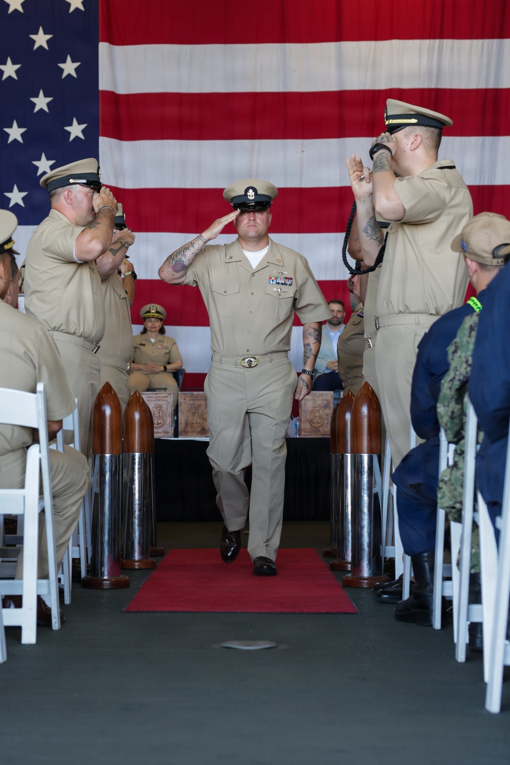 A U.S. Navy military ceremony is taking place indoors, with a central figure in khaki service uniform walking down a red carpet while being saluted by two flanking personnel. A large American flag serves as a backdrop, and decorative artillery shell replicas line the aisle. The setting and attire suggest a formal event such as a chief petty officer pinning ceremony or retirement/promotion ceremony.