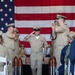 A U.S. Navy military ceremony is taking place indoors, with a central figure in khaki service uniform walking down a red carpet while being saluted by two flanking personnel. A large American flag serves as a backdrop, and decorative artillery shell replicas line the aisle. The setting and attire suggest a formal event such as a chief petty officer pinning ceremony or retirement/promotion ceremony.