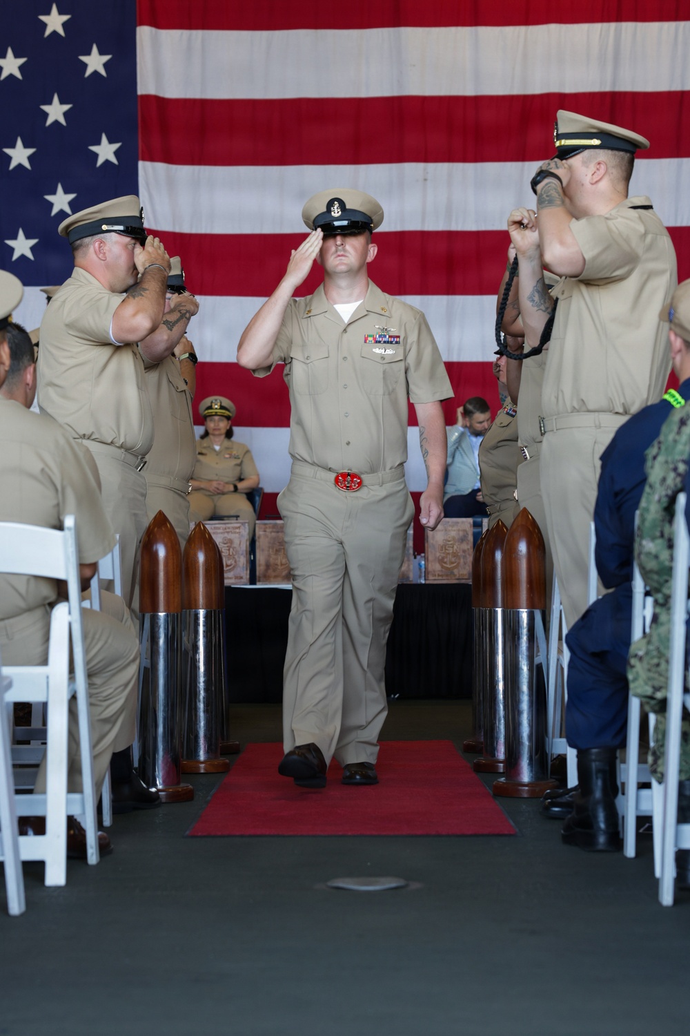 A U.S. Navy Chief Petty Officer initiation or pinning ceremony is taking place in what appears to be a hangar bay or large indoor venue. The central figure, dressed in khaki service uniform, is saluting while walking down a red carpet flanked by two ceremonial shell casings and fellow sailors rendering salutes. A large American flag serves as the backdrop, and seated attendees in various uniforms observe the proceedings.