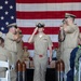 A U.S. Navy Chief Petty Officer initiation or pinning ceremony is taking place in what appears to be a hangar bay or large indoor venue. The central figure, dressed in khaki service uniform, is saluting while walking down a red carpet flanked by two ceremonial shell casings and fellow sailors rendering salutes. A large American flag serves as the backdrop, and seated attendees in various uniforms observe the proceedings.