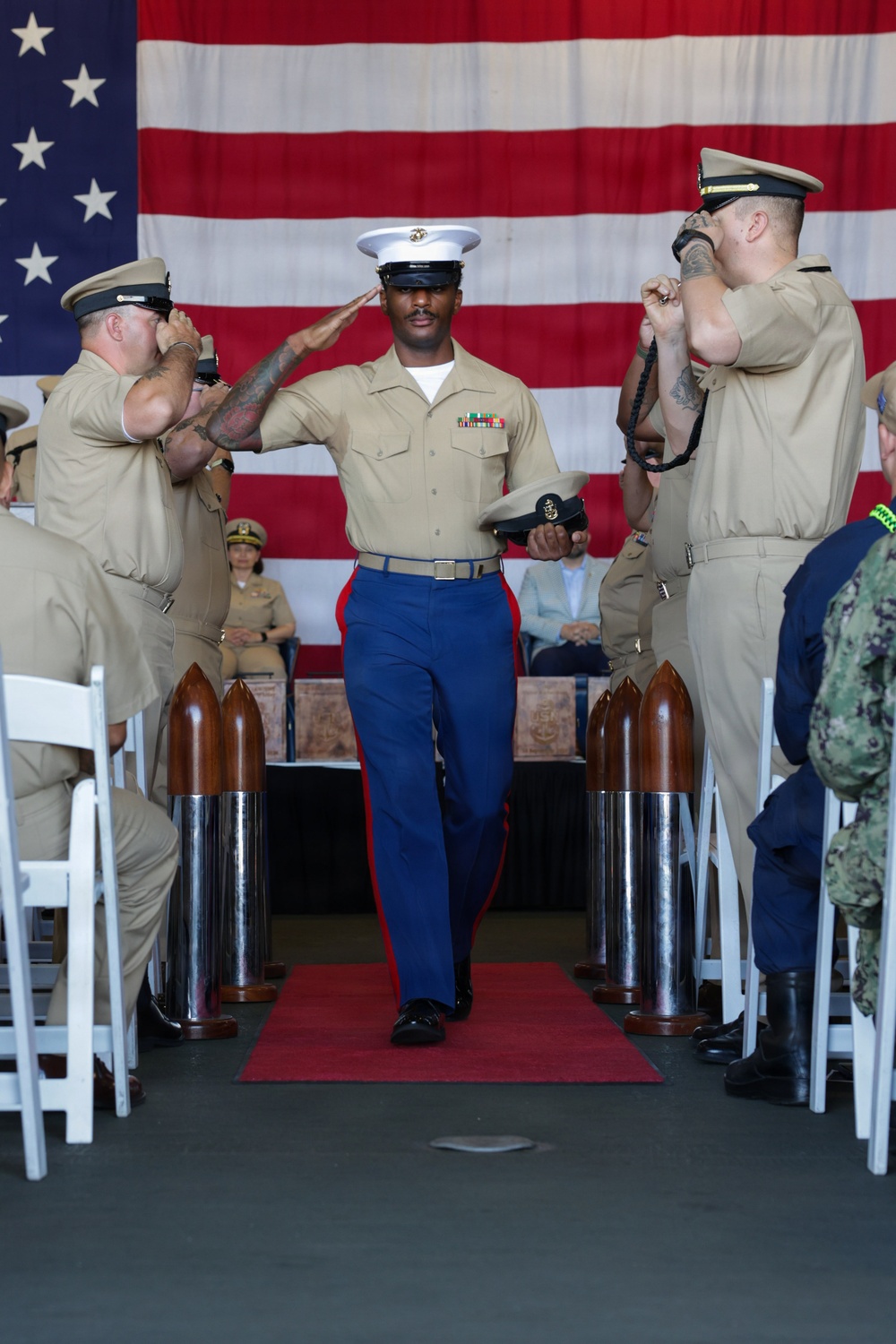 A U.S. Marine in dress uniform walks down a red carpet aisle during a formal military ceremony, rendering a salute while flanked by Navy personnel in khaki uniforms who are also saluting. The ceremony takes place in front of a large American flag backdrop, with decorative artillery shell replicas lining the aisle. The Marine is holding a cover (hat) in his left hand while saluting with his right, suggesting a promotion or retirement ceremony.