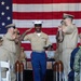 A U.S. Marine in dress uniform walks down a red carpet aisle during a formal military ceremony, rendering a salute while flanked by Navy personnel in khaki uniforms who are also saluting. The ceremony takes place in front of a large American flag backdrop, with decorative artillery shell replicas lining the aisle. The Marine is holding a cover (hat) in his left hand while saluting with his right, suggesting a promotion or retirement ceremony.