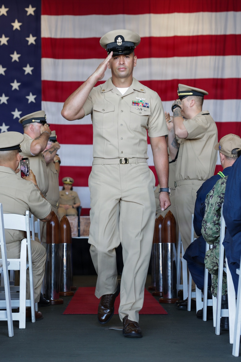 A U.S. Navy Chief Petty Officer in Service Khaki uniform walks down a ceremonial aisle while rendering a salute, with an American flag displayed prominently in the background. The setting appears to be a formal military ceremony, likely a Chief Petty Officer pinning or promotion ceremony, with rows of seated Navy personnel on both sides. Several attendees are photographing the event on smartphones.