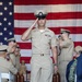 A U.S. Navy Chief Petty Officer in Service Khaki uniform walks down a ceremonial aisle while rendering a salute, with an American flag displayed prominently in the background. The setting appears to be a formal military ceremony, likely a Chief Petty Officer pinning or promotion ceremony, with rows of seated Navy personnel on both sides. Several attendees are photographing the event on smartphones.