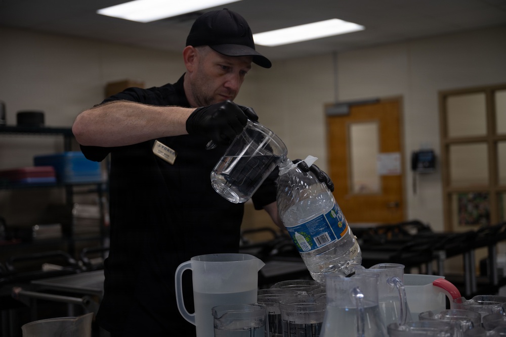 A man dressed in all black, wearing a black cap and black gloves, is pouring water from a large plastic water bottle into a clear glass pitcher. He appears to be in an institutional or commercial setting, surrounded by multiple plastic and glass pitchers on a table. A name badge is visible on his chest, suggesting a professional or service role.