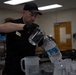 A man dressed in all black, wearing a black cap and black gloves, is pouring water from a large plastic water bottle into a clear glass pitcher. He appears to be in an institutional or commercial setting, surrounded by multiple plastic and glass pitchers on a table. A name badge is visible on his chest, suggesting a professional or service role.